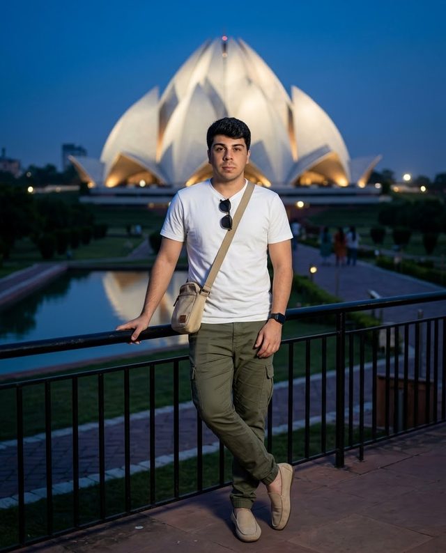 Flávio Vieira, de camiseta branca e calça verde-oliva, apoiado em um gradil ao anoitecer, com o Templo de Lótus iluminado ao fundo.