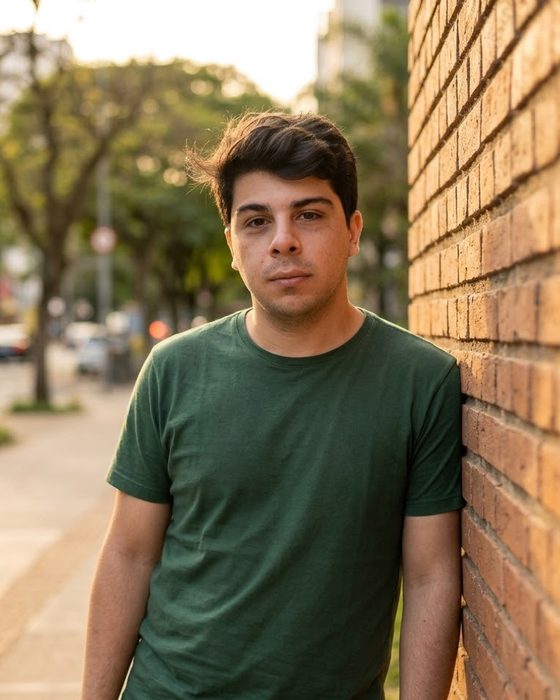 Flávio A. Vieira, de camiseta verde, encostado em um muro de tijolos numa calçada arborizada, no fim da tarde.